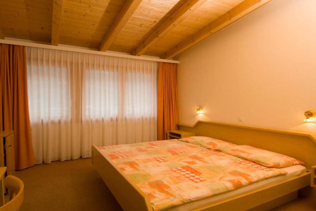 A bedroom with a wooden ceiling, a bed with a patterned bedspread, a nightstand, a desk, and a chair.