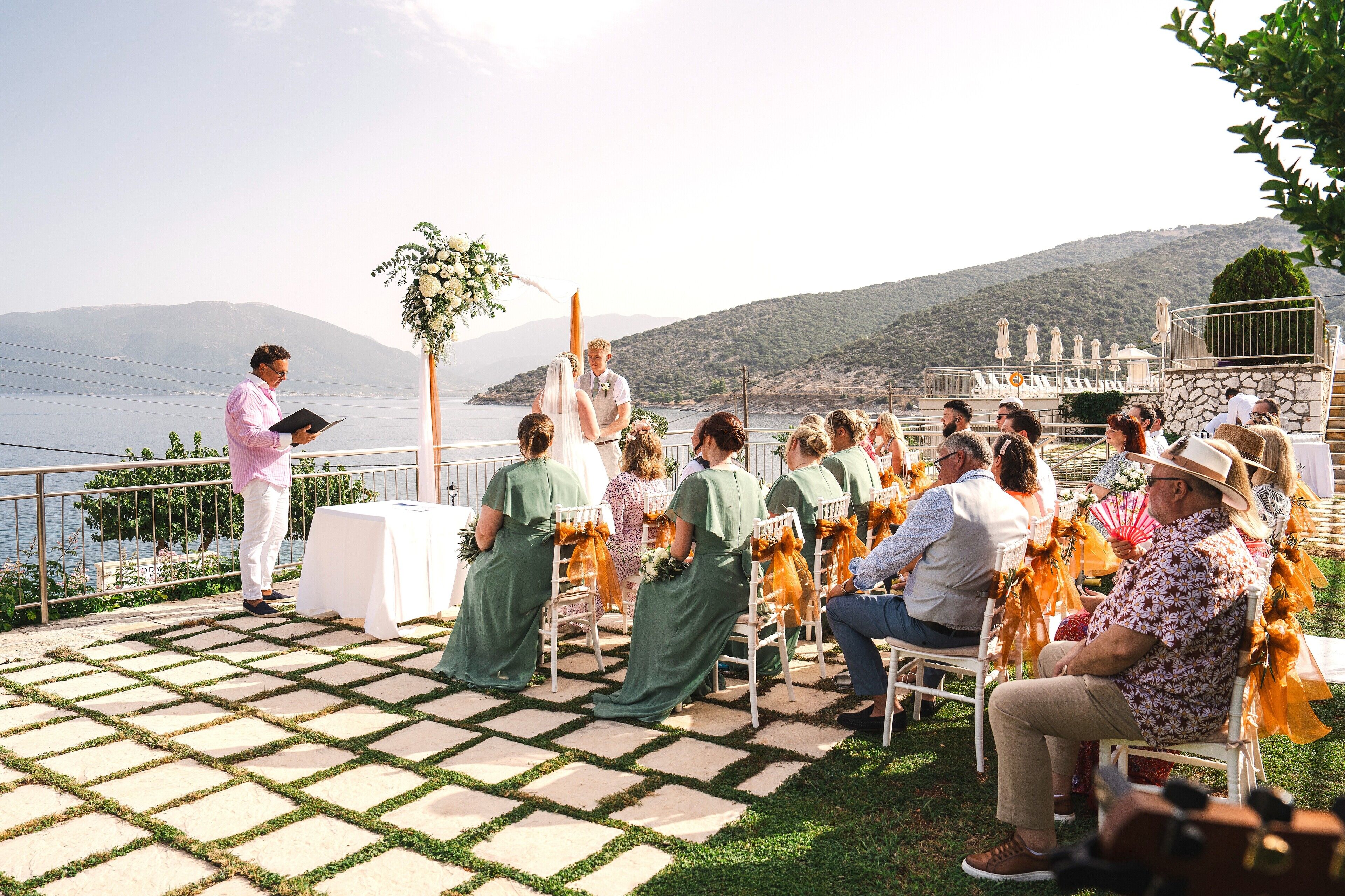 A wedding ceremony outdoors with guests seated on chairs, a speaker at the front, and a scenic backdrop of hills and water.
