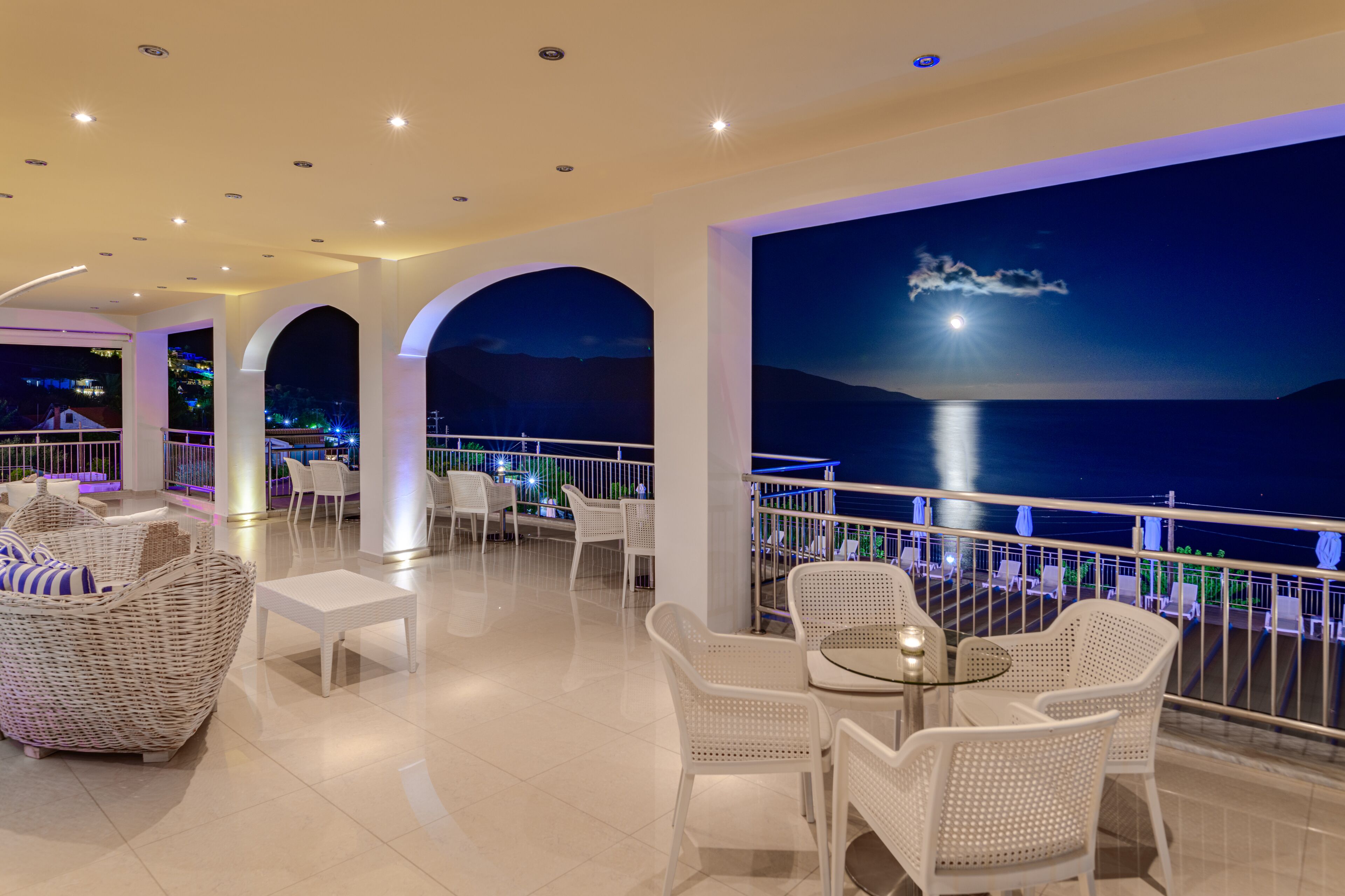 A spacious balcony with white wicker furniture and a view of the sea at night.