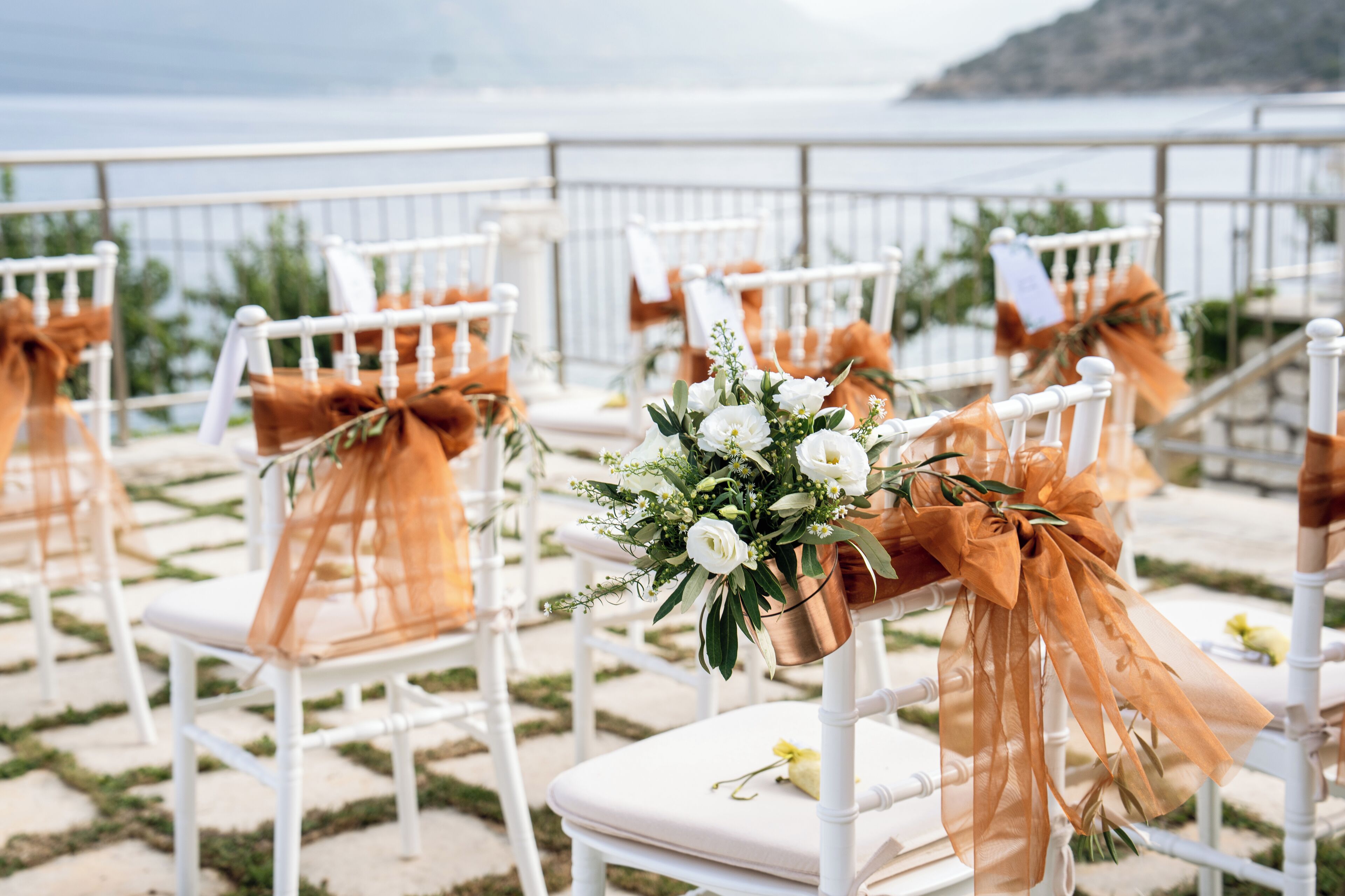 Rows of white chairs with orange sashes and floral arrangements, set up on a terrace with a view of the sea.