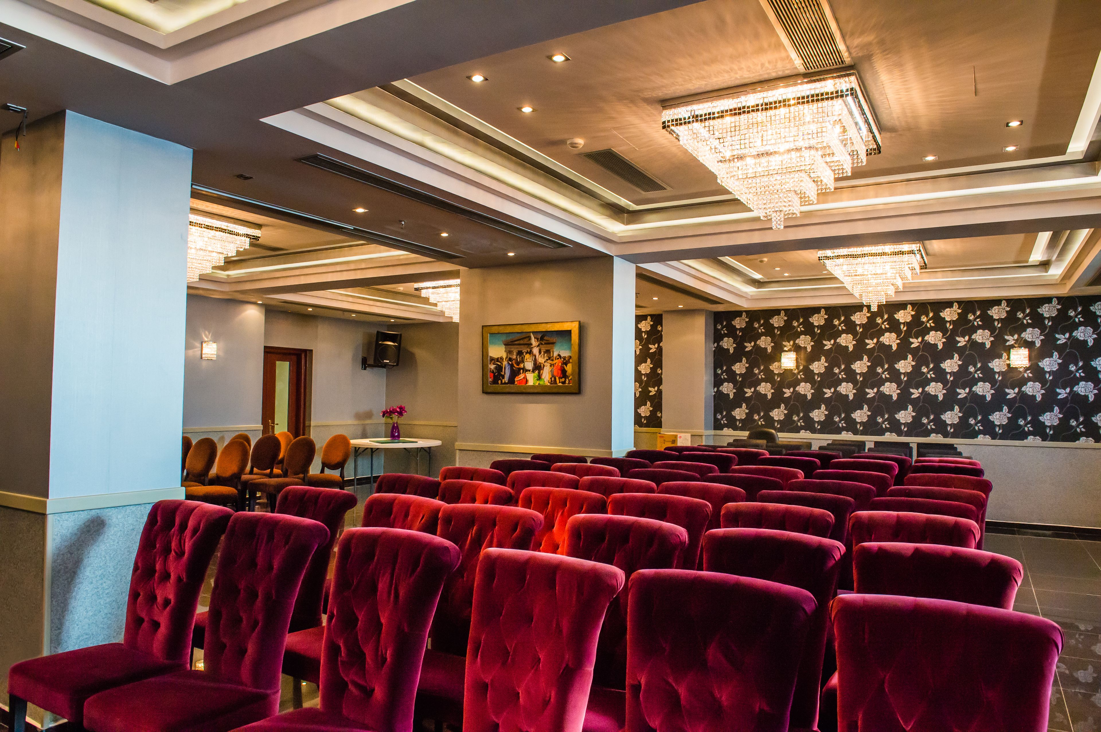 A conference room with rows of red velvet chairs, a chandelier, and a floral wallpapered wall.