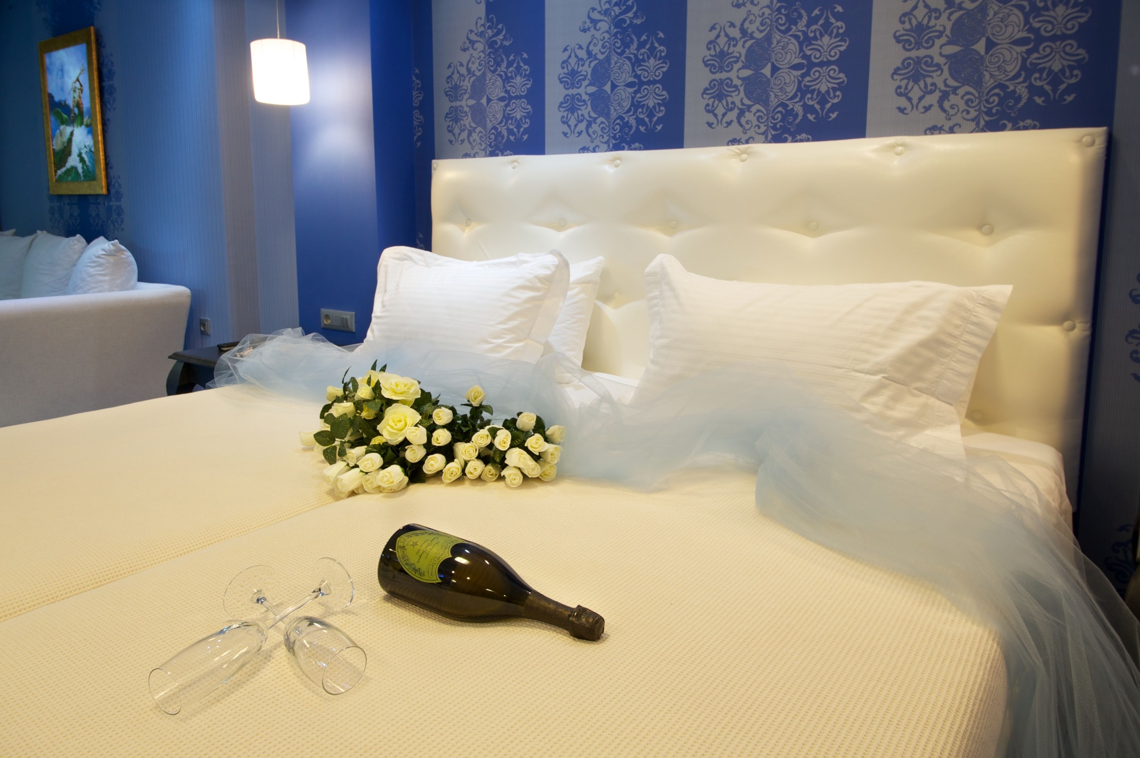Una cama bien hecha con sábanas blancas, un ramo de rosas blancas, una botella de champán y un par de copas de vino sobre una colcha blanca con textura.