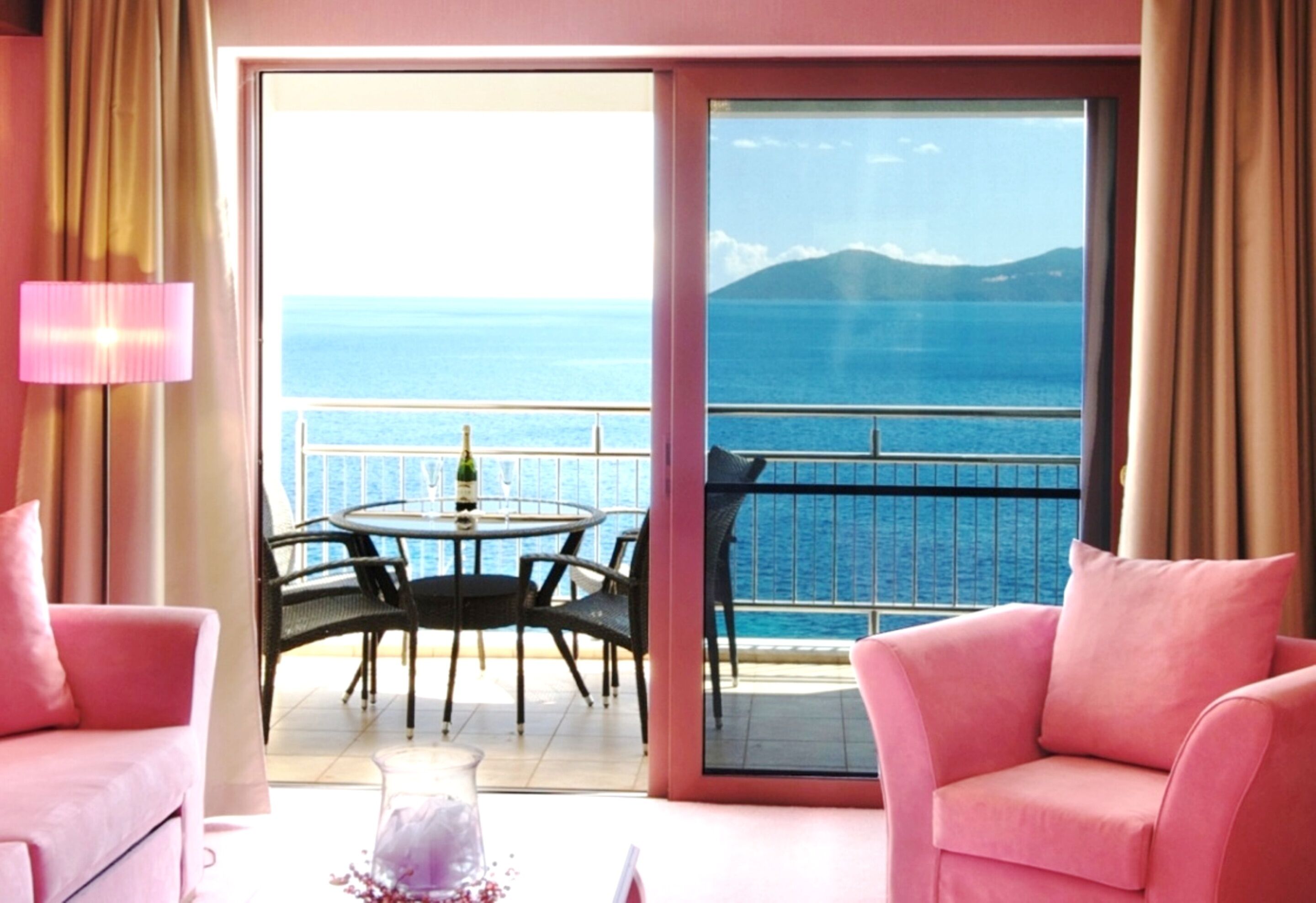 A room with a balcony, pink furniture, a table with chairs, and a view of the sea and mountains.