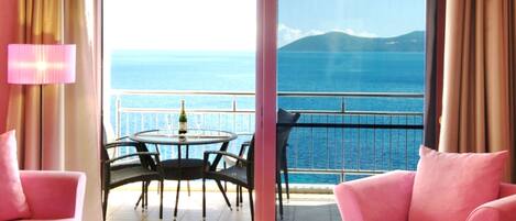 A room with a balcony, pink furniture, a table with chairs, and a view of the sea and mountains.