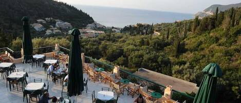Une terrasse extérieure, agréée de tables et de chaises, qui offre une vue sur un paysage côtier avec des collines et la mer.