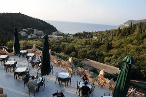 An outdoor terrace with tables and chairs, overlooking a coastal landscape with hills and the sea.