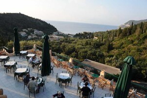 Una terraza exterior con mesas y sillas, con vistas a un paisaje costero con colinas y el mar.