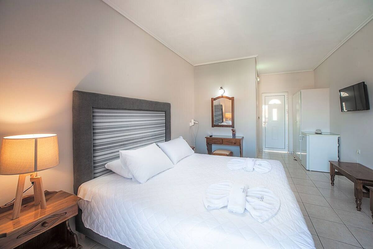 A hotel room with a large bed, a bedside table with a lamp, a dresser, a mirror, a television, and a refrigerator.