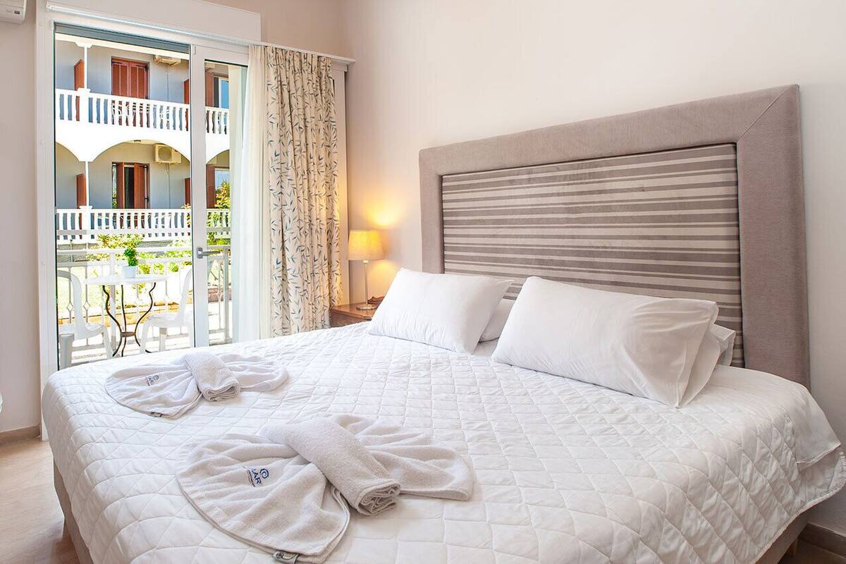 A hotel room with a bed, a balcony with a white railing, and a view of the outside.