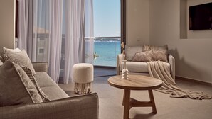 A modern living room with a sofa, armchair, and ottoman, a round wooden table, and a view of the sea through a large window. - Windmill Bay Hotel (Zakynthos)