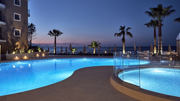 A swimming pool with a curved edge, surrounded by lounge chairs and palm trees, with a view of the ocean and a building in the background. - Windmill Bay Hotel (Zakynthos)