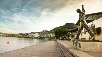 A wooden boardwalk with a statue, overlooking a river with buildings and hills in the background.
