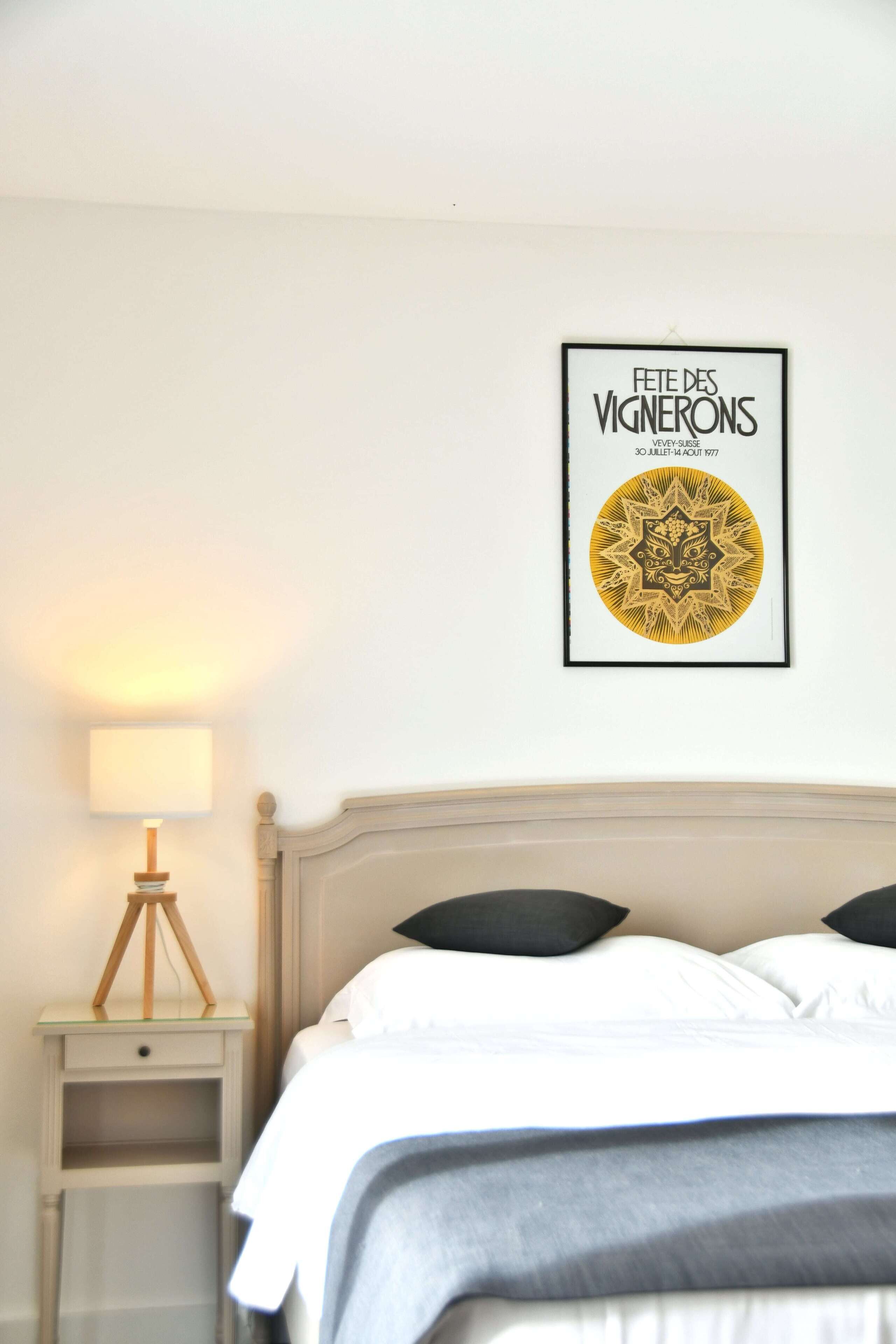 Un lit bien fait, avec une literie blanche et grise, une œuvre d’art encadrée au mur et une lampe de chevet sur une table de nuit.
