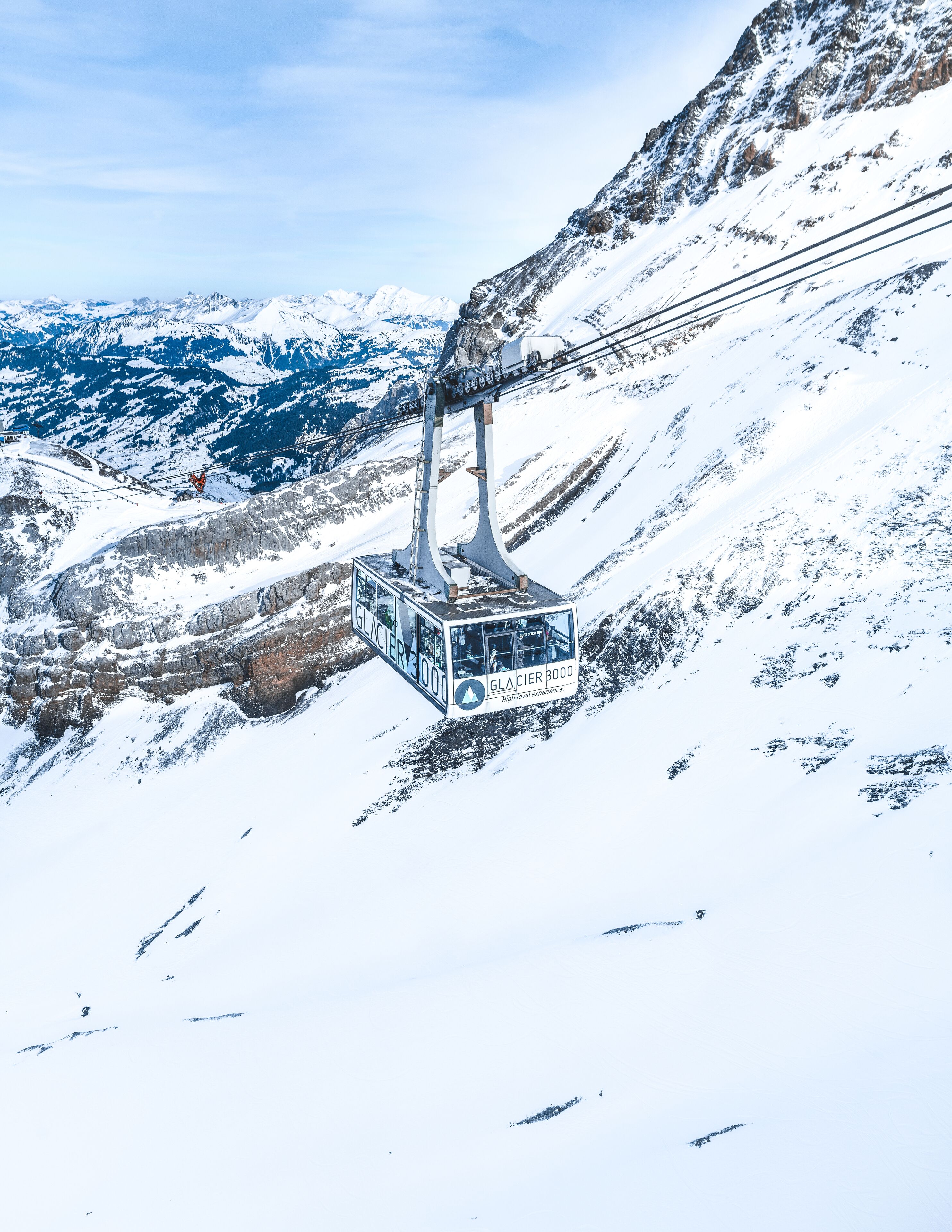 Un teleférico con la etiqueta "GLACIER 3000" suspendido sobre un paisaje montañoso nevado.
