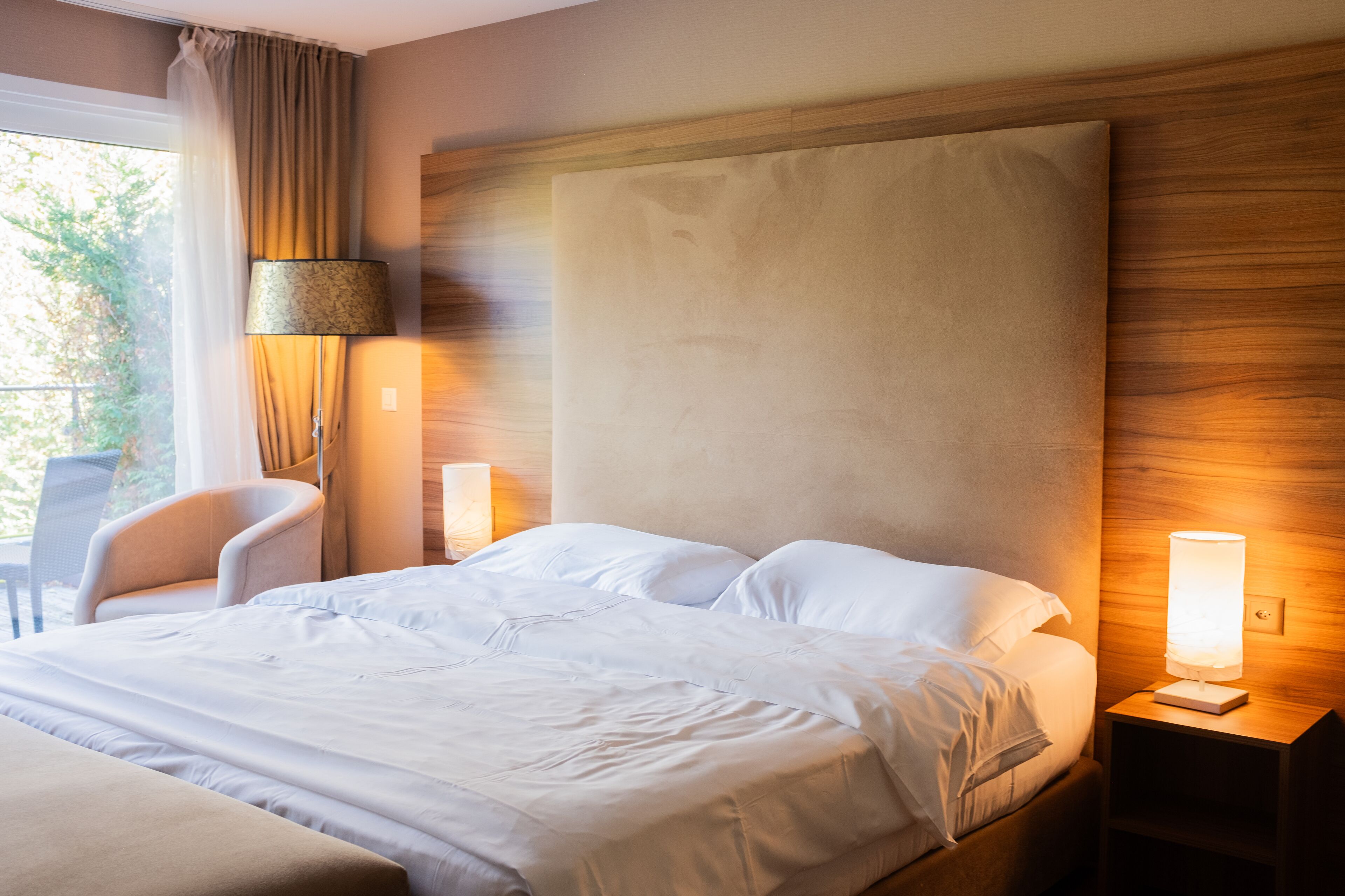 Habitación de hotel con una cama grande, una silla, una mesita de noche con lámpara y una ventana con cortinas.