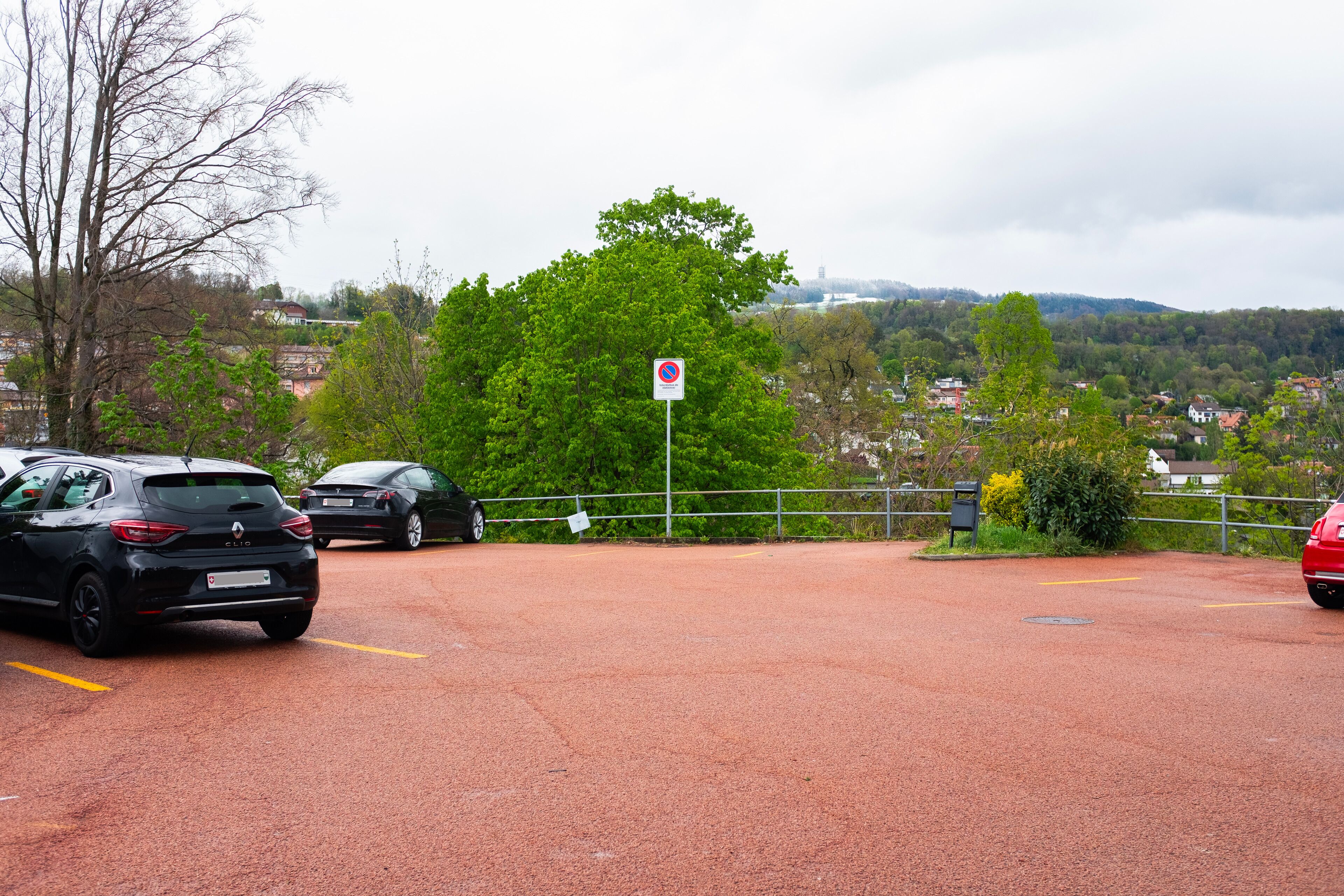 Un parking rempli de voitures, un panneau indiquant l’interdiction de se garer, et en arrière-plan, une vue sur des maisons et des arbres.