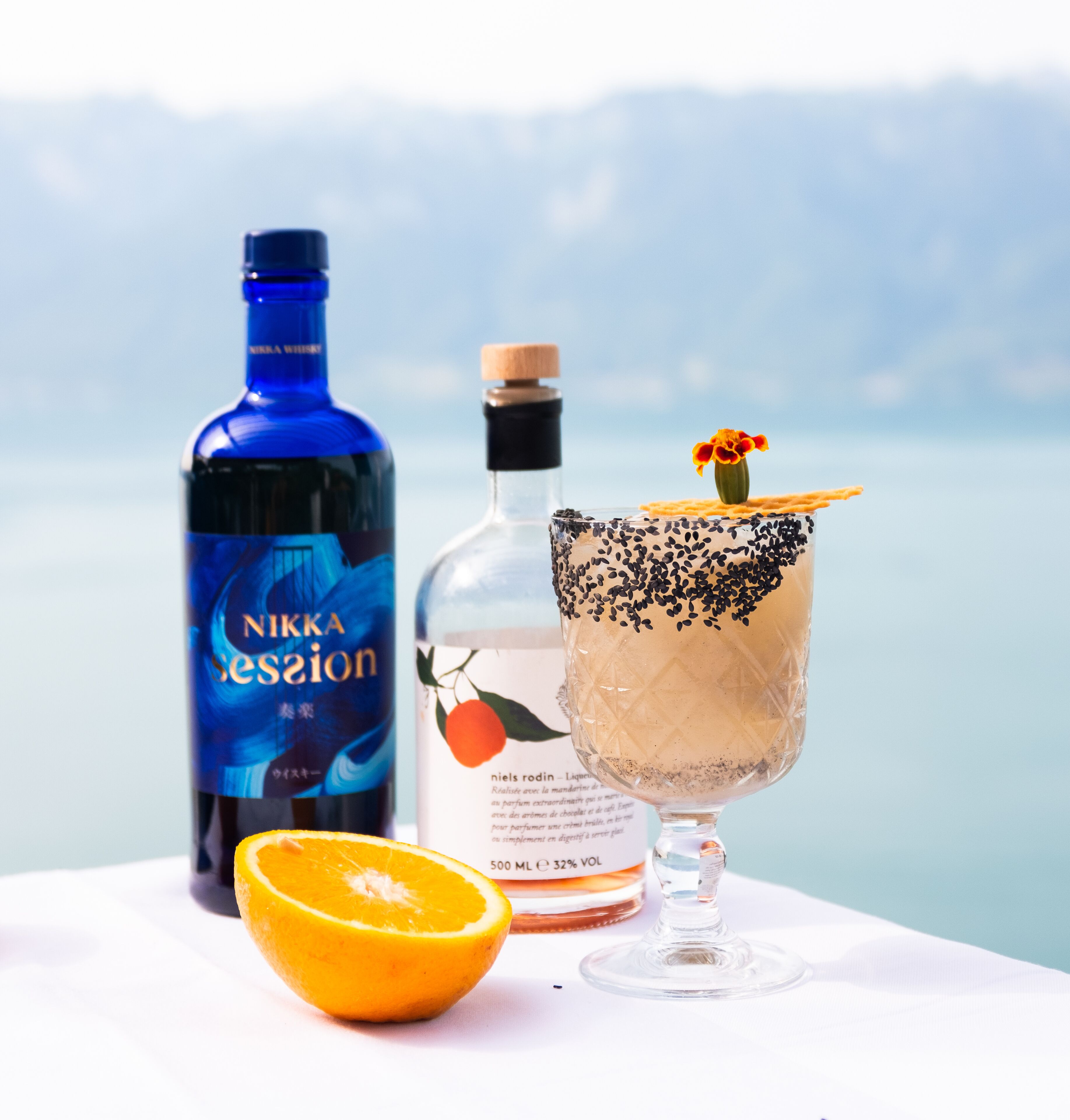 Una botella de whisky Nikka, una copa con un cóctel decorado con semillas negras y una flor, y una rodaja de naranja sobre una mesa con una vista panorámica al fondo.