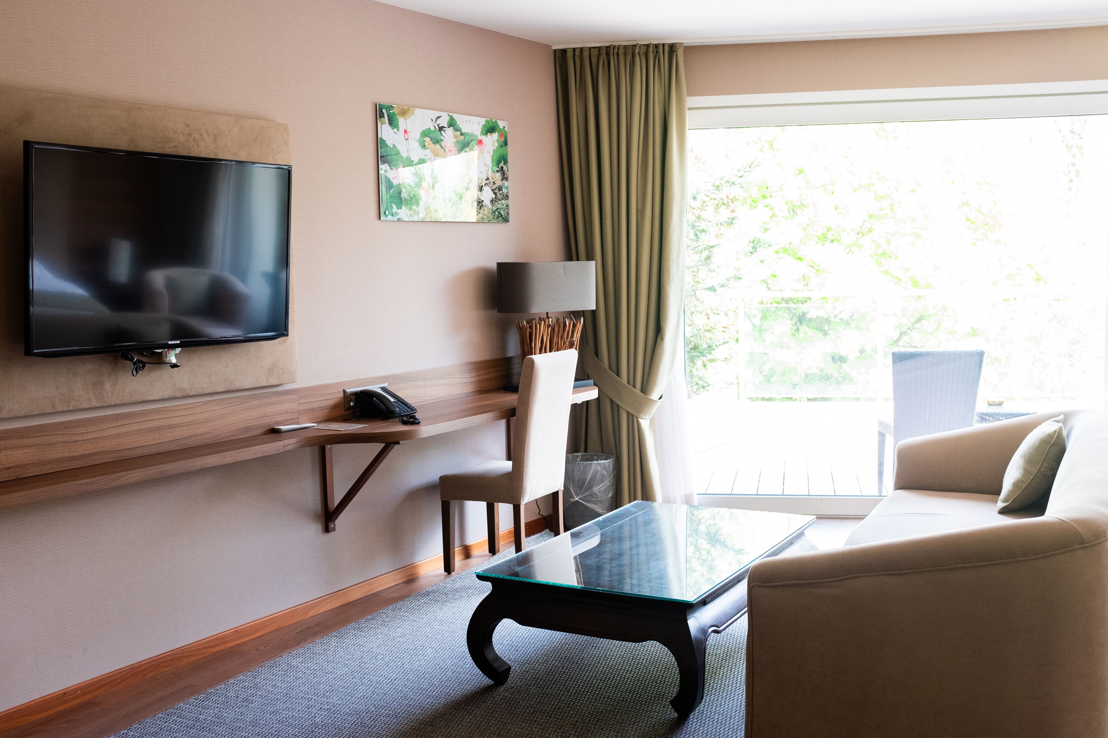 Quarto de hotel com televisão de tela plana na parede, mesa de centro de vidro, sofá bege, uma cadeira e uma janela com cortinas.