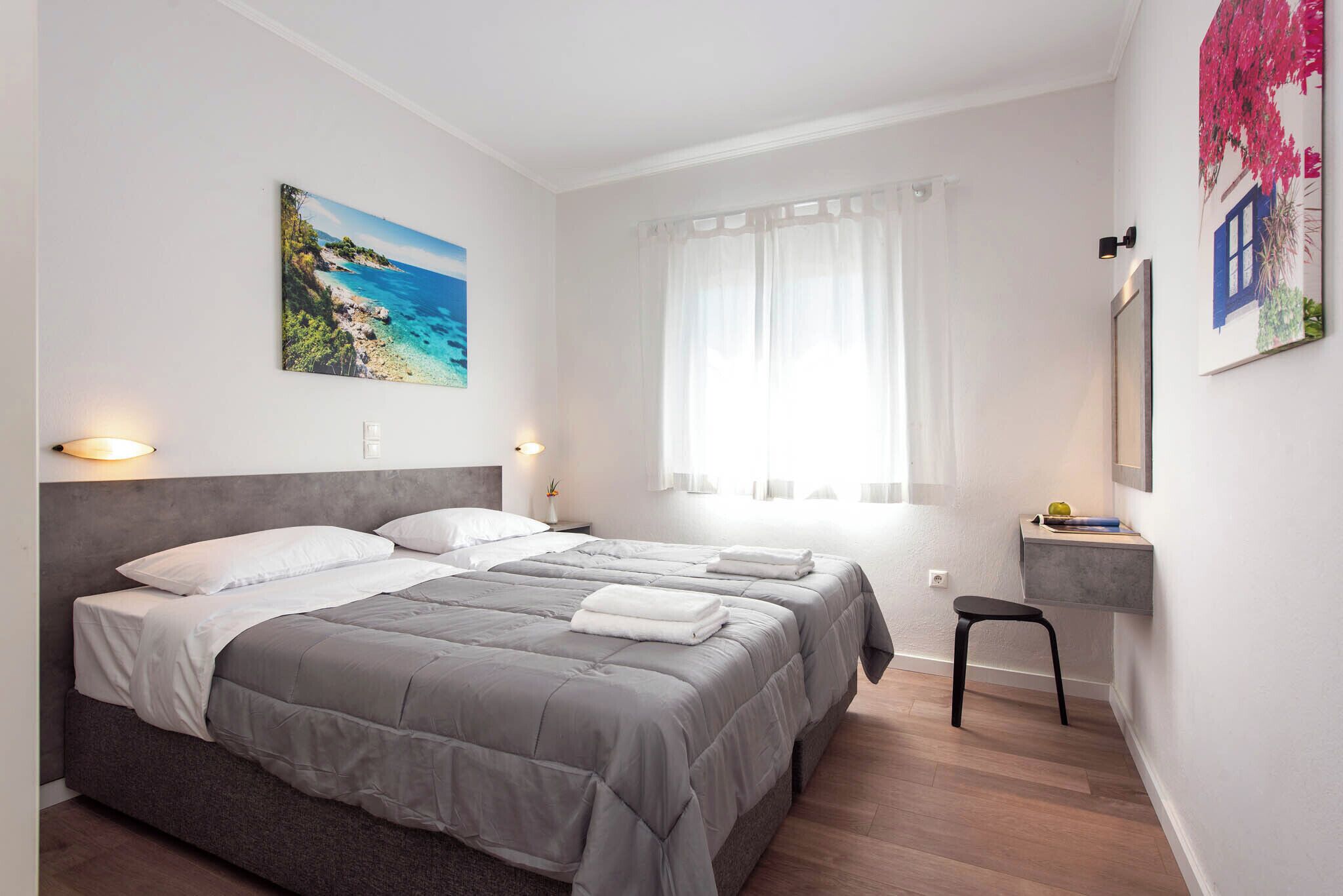 A bedroom with two beds, a window with sheer curtains, a painting of a beach, and a small desk with a chair.