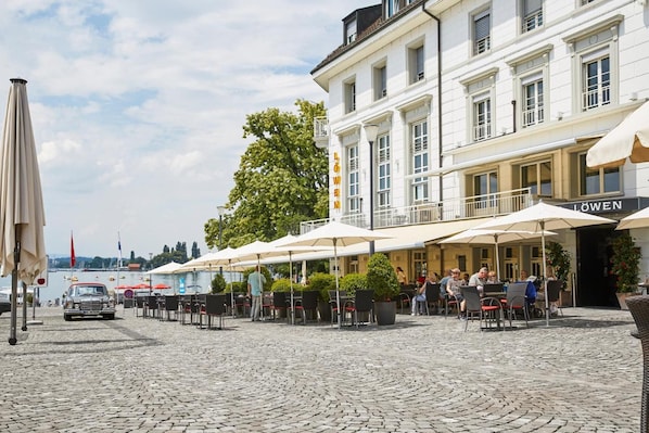 A cobblestone courtyard with outdoor seating and umbrellas, a building with 'LOWEN' signage, and a parked car. - Hotel Löwen Am See (Zug)