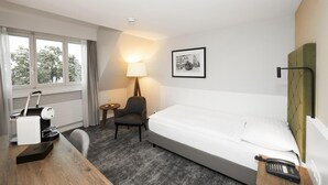 Una habitación de hotel con una cama, un escritorio con cafetera, una silla, una lámpara y un cuadro en la pared.