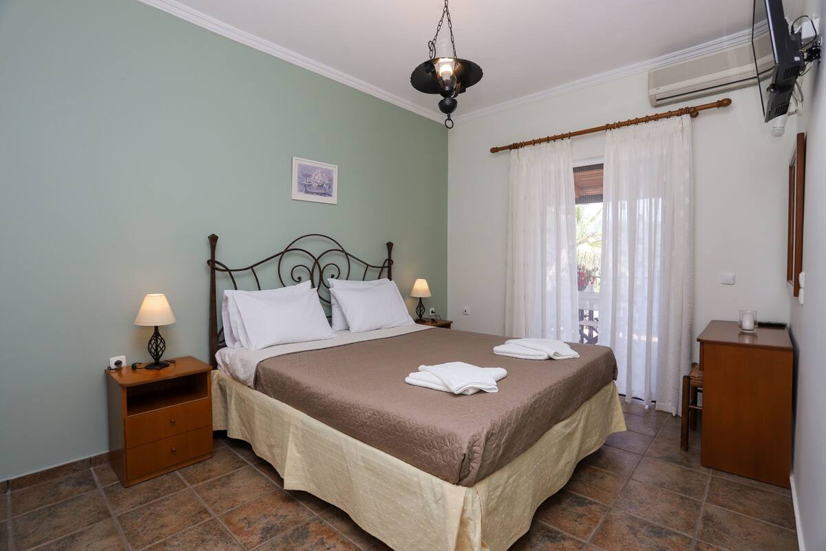 Una cama bien hecha con una colcha marrón y almohadas blancas, una mesita de noche con una lámpara y una mesita auxiliar de madera.