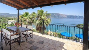 Uma área de estar coberta ao ar livre, com mesa e cadeiras, com vista para a costa, palmeiras e mar azul cristalino.