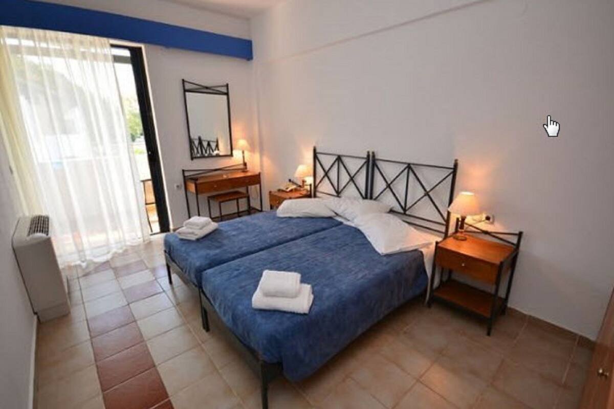 A double bed with a blue bedspread, two bedside tables with lamps, a desk with a chair, a large mirror, and a sliding glass door leading to a balcony.