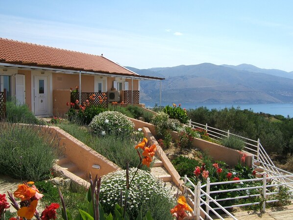 Une maison avec un toit recouvert de tuiles rouges, un balcon et un jardin où poussent différentes sortes de fleurs.