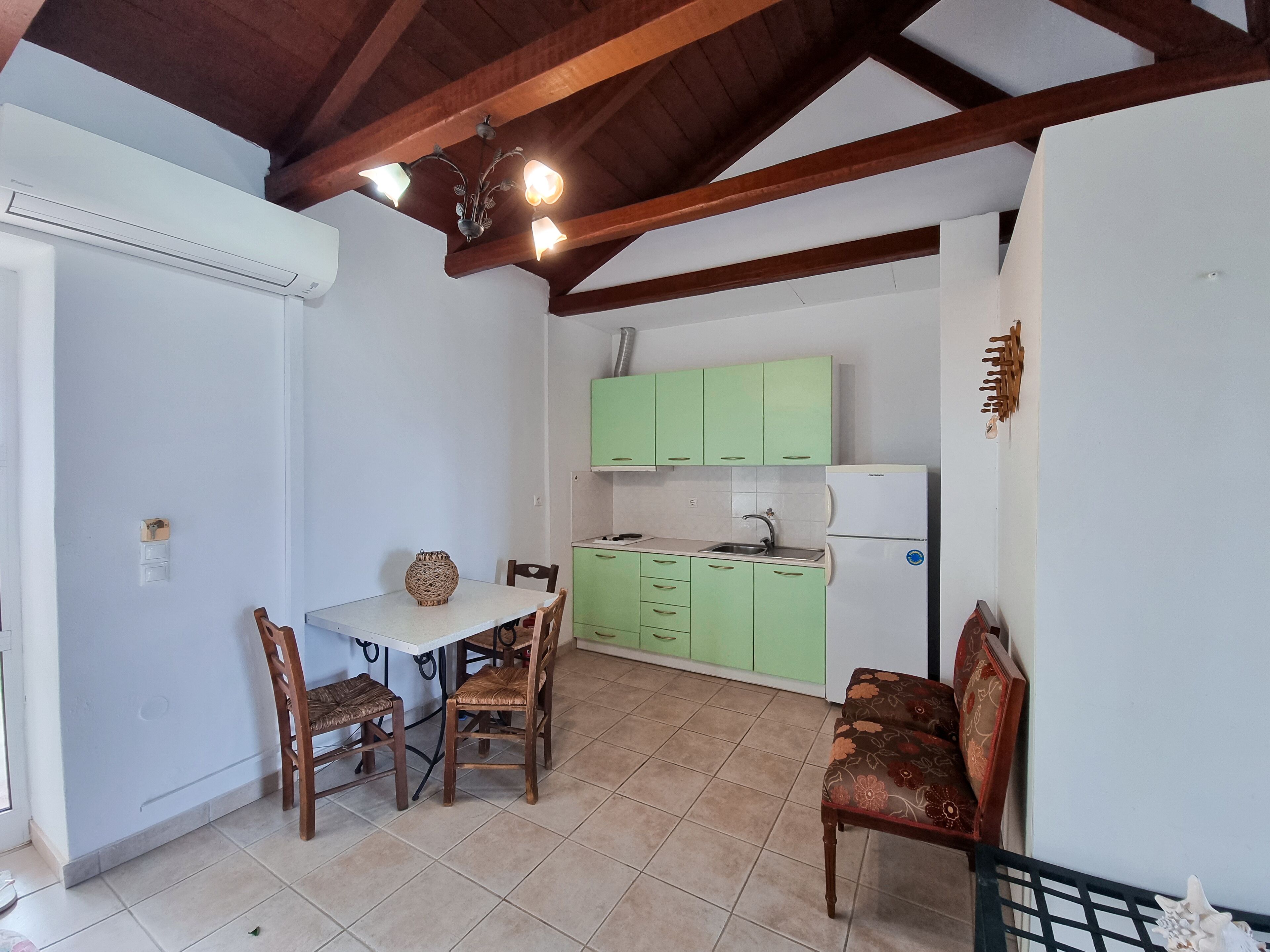Cocina compacta con armarios verdes, un refrigerador blanco y una zona de comedor con dos sillas de madera y una mesa.