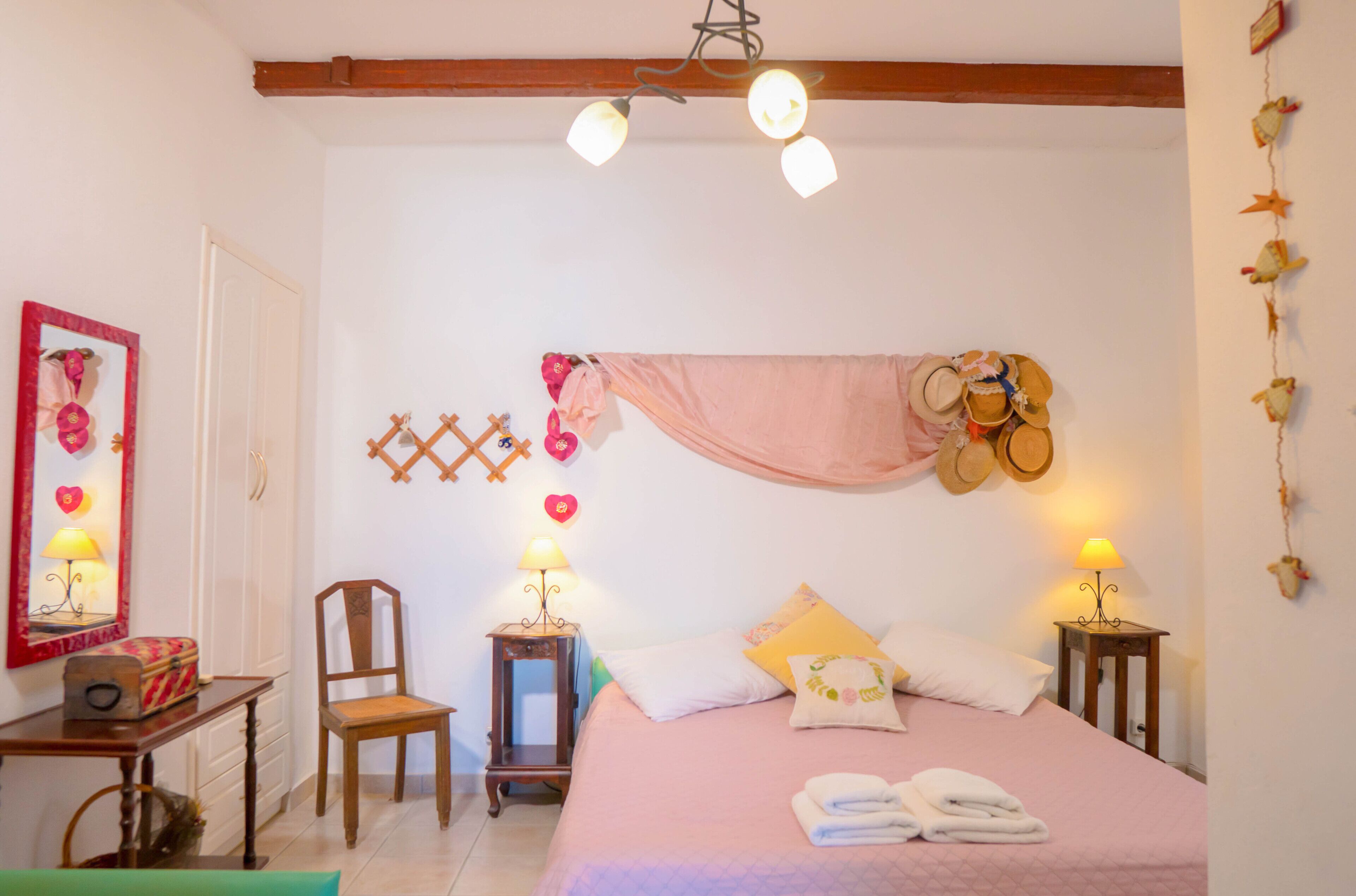 A bedroom with a bed, two bedside tables with lamps, a chair, a wall-mounted rack with hats, and a decorative mirror.