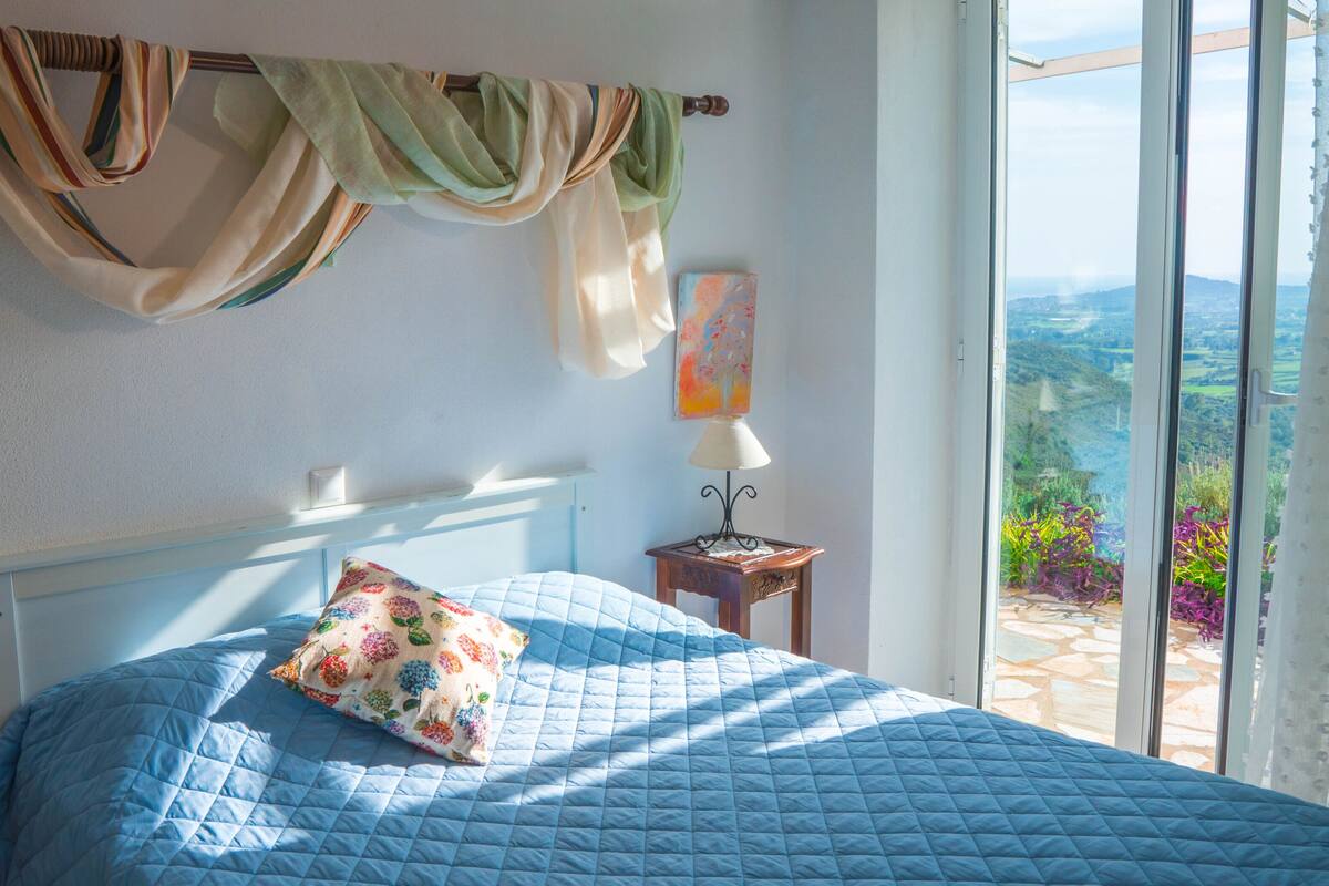 Un dormitorio con cama, mesita de noche, lámpara, ventana con vistas y cortina.