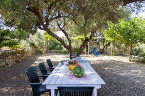 An outdoor dining area with a white table, black chairs, and a basket of fruits. - Forestata Village (Kefalonia)