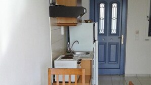A compact kitchen with a blue door, a wooden table with a fruit bowl, and a small chair. - Agnanti Studios (Kefalonia)