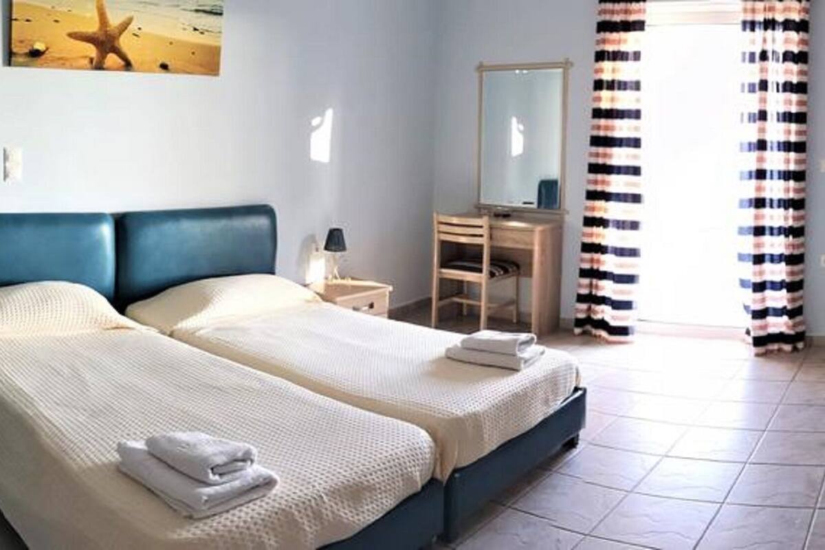 Un lit double avec une tête de lit bleue, une table de chevet avec une lampe, une commode, un miroir, des rideaux à rayures et une petite table avec un vase.