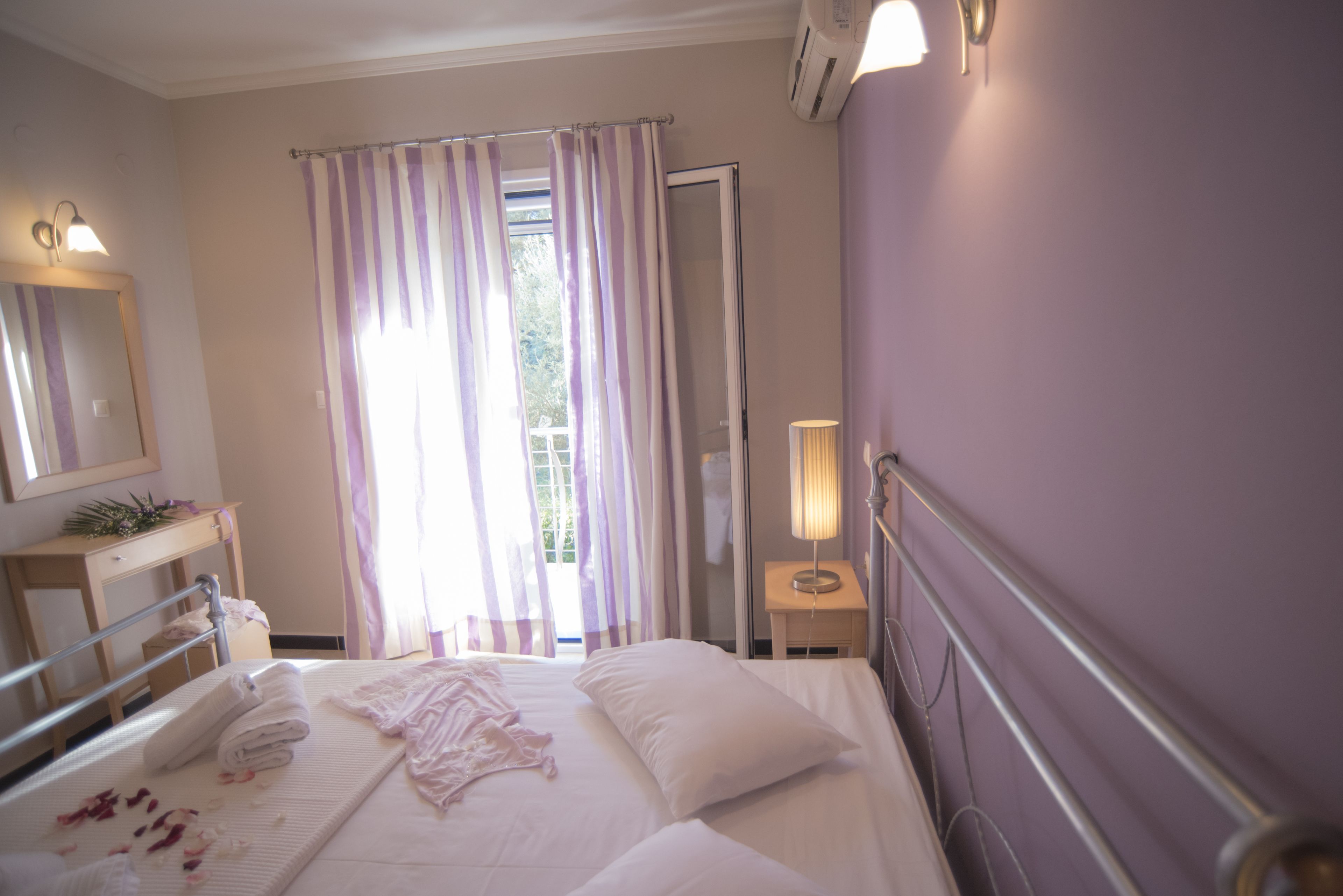 A neatly made bed with pillows and a folded towel, a bedside table with a lamp, a window with curtains, and a mirror on the wall.