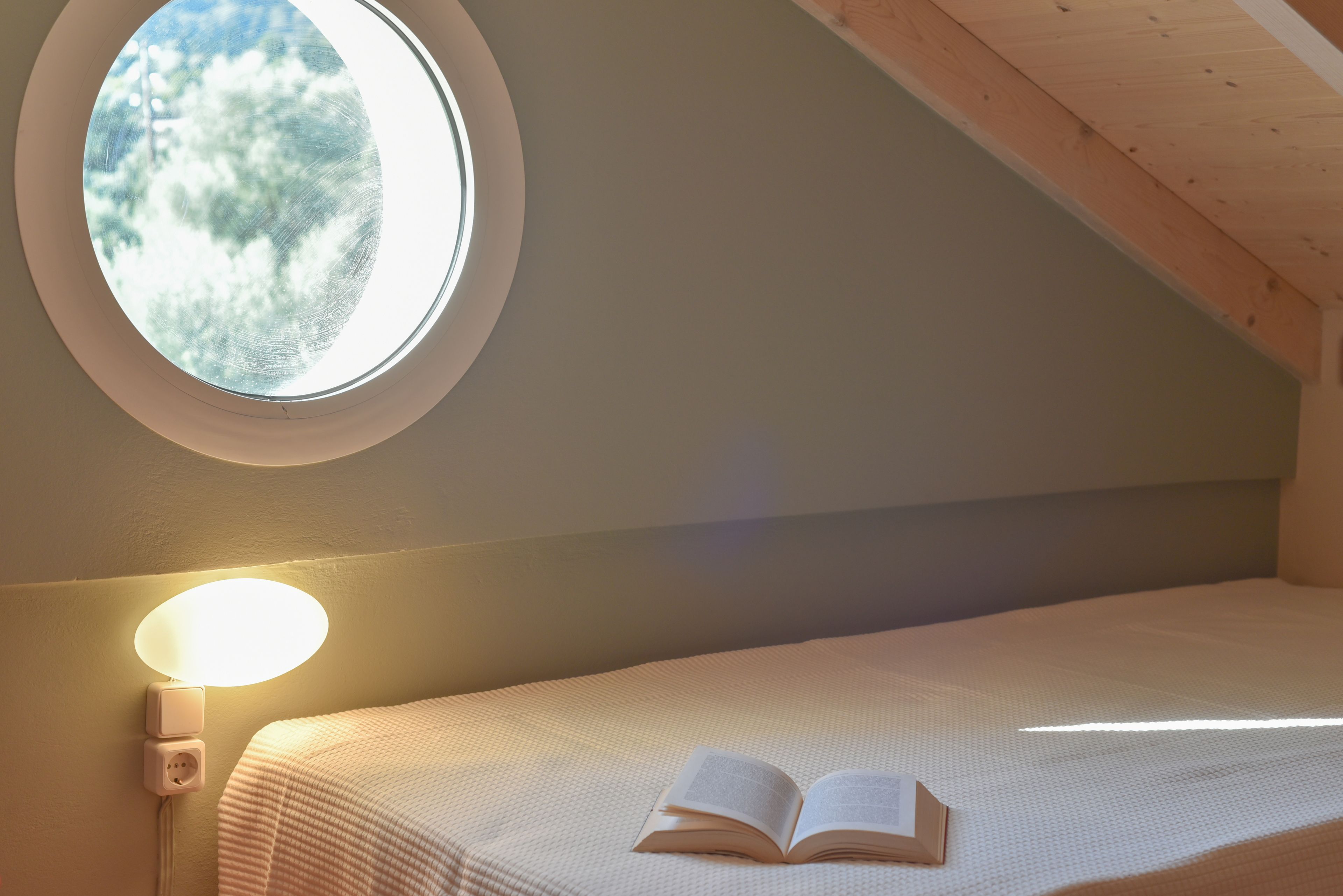 Un dormitorio con tragaluz, una lámpara de noche y un libro abierto sobre la cama.