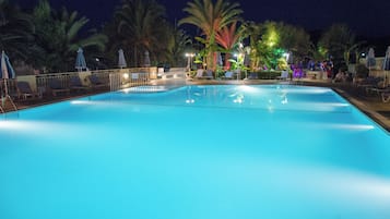 A nighttime swimming pool with lounge chairs and umbrellas, surrounded by palm trees and illuminated by ambient lighting.
