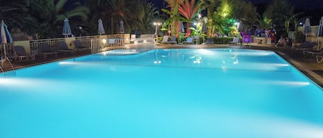 Una piscina con sillas de descanso y sombrillas, rodeada de palmeras e iluminada por luces nocturnas.