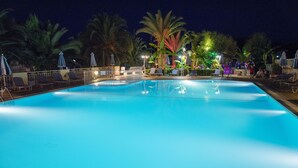 Una piscina iluminada con luces ambientales, sillas de descanso y sombrillas, rodeada de palmeras.