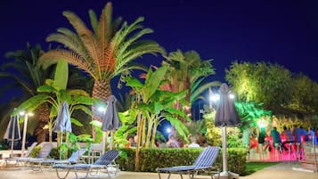 A nighttime outdoor area with lounge chairs and umbrellas, surrounded by palm trees and greenery.