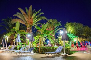 A nighttime outdoor area with lounge chairs and umbrellas, surrounded by palm trees and greenery. - Athos Hotel (Lefkada)