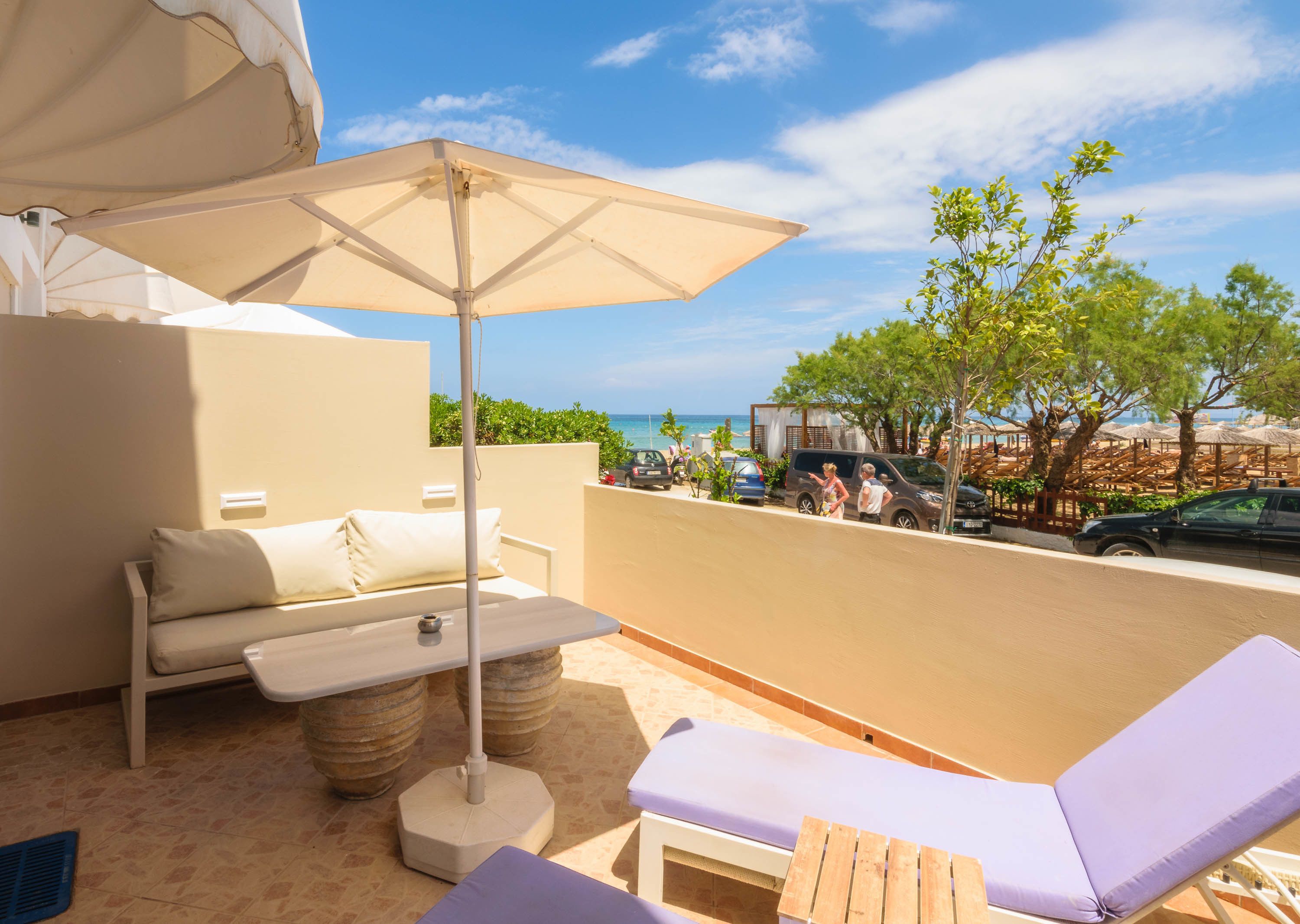 A balcony with a sofa, a table, and a lounge chair, overlooking a beach with umbrellas and people.