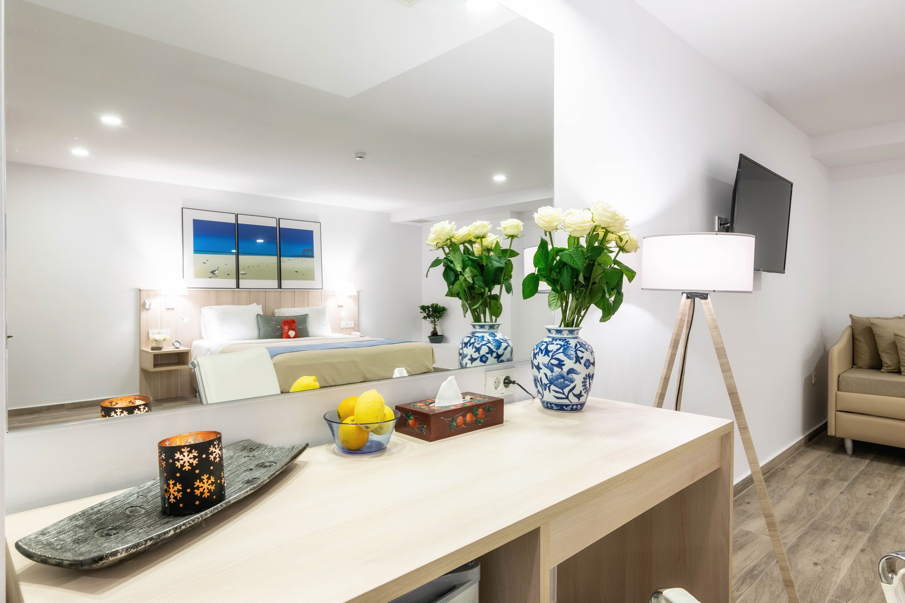 Ett modernt hotellrum med en stor säng, ett sängbord med en lampa, en spegel, en tv och en vas med blommor.