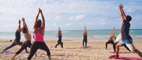 Un grupo de personas practicando yoga sobre esterillas en una playa arenosa, con el océano de fondo.