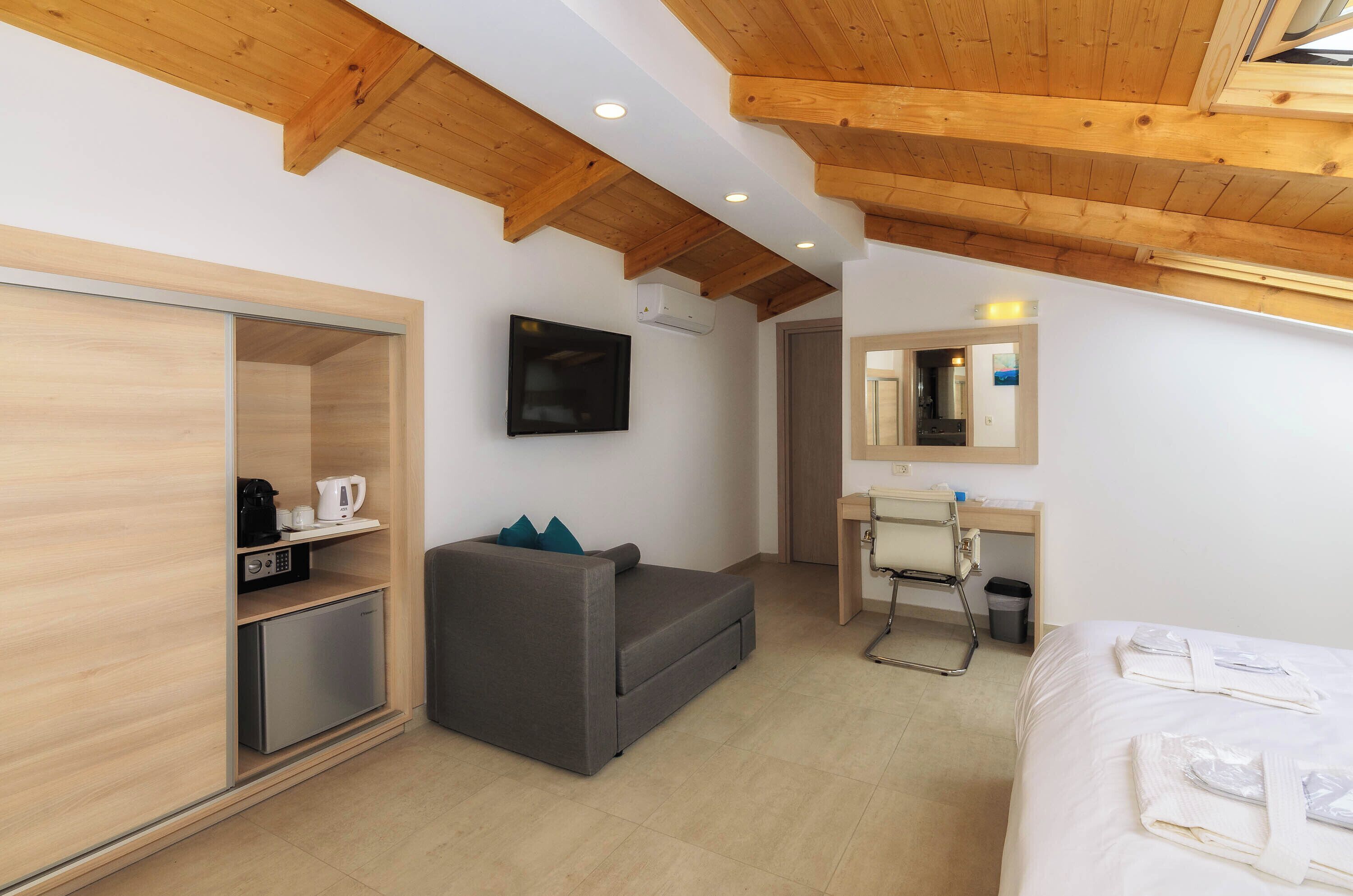 A modern hotel room with a wooden ceiling, a grey sofa, a small desk with a chair, a bed with neatly folded towels, and a television on the wall.