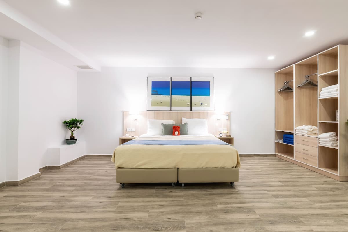 A hotel room with a bed, a wooden wardrobe, a nightstand, a potted plant, and a painting of a beach scene.