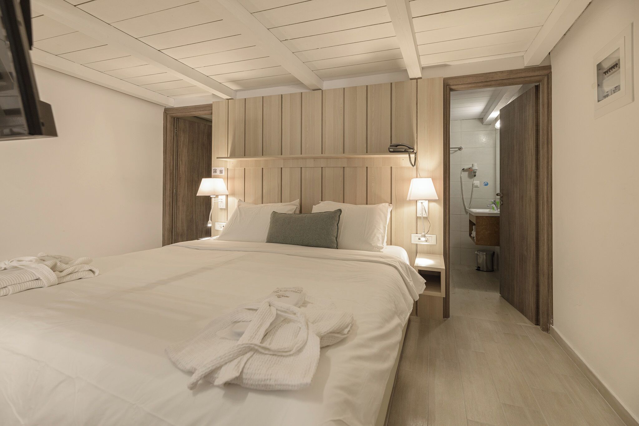 Una camera d'albergo con un letto grande, due lampade ai lati del letto, una testiera in legno, un bagno visibile attraverso una porta aperta e un asciugamano sul letto.