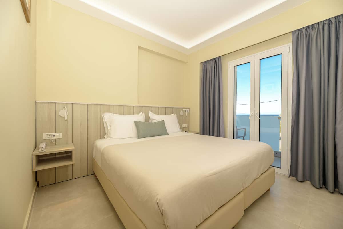 A hotel room with a large bed, a bedside table, a power outlet, and a view through a sliding door to the outside.