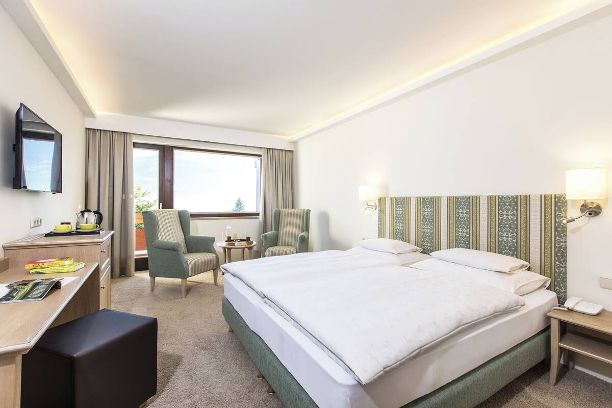 A hotel room with a large bed, a desk with a chair, a TV mounted on the wall, and a window with curtains.